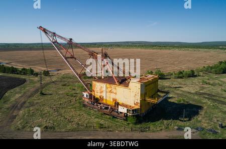 L'enorme escavatore dragline industriale poggia in un campo aperto, circondato da vegetazione sparsa e strade sterrate Foto Stock