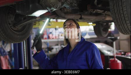 Ispezione di una donna meccanica in tute blu con luce a LED, esame della parte inferiore dell'ascensore in officina Foto Stock