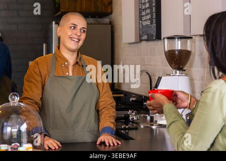 Barista donna in grembiule verde e cliente donna che serve una tazza rossa, chiacchierando dietro il bancone del bar Foto Stock
