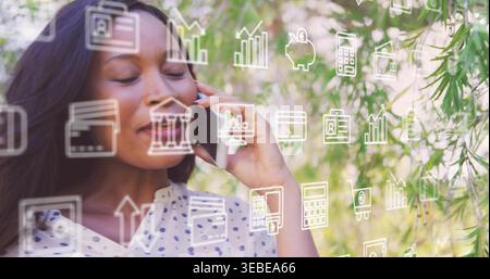 Donna in piedi che indossa una camicetta bianca punteggiata che tiene il telefono all'orecchio nel parco, mostrando icone finanziarie Foto Stock