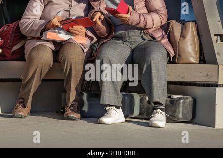Due amici anziani si siedono sulla panchina all'aperto condividendo i contenuti dello smartphone con copertina ribaltabile e scambiando foto mentre si riscaldano alla luce del sole nel tardo pomeriggio. Foto Stock
