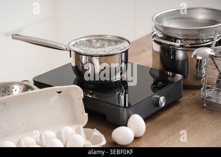 Pentola con acqua bollente su un piccolo piano cottura e uova in cucina Foto Stock