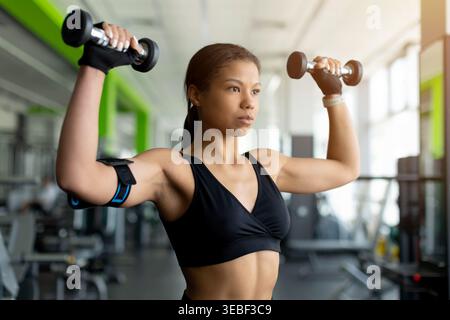 Giovane donna nera concentrata che fa una pressa per spalla con manubri in palestra Foto Stock