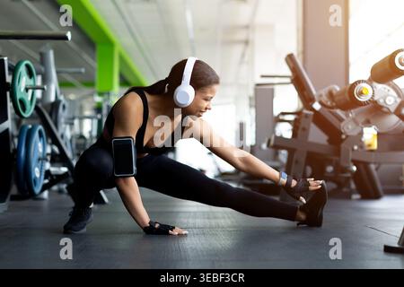 La giovane donna nera che fa un affondo laterale si allunga sul pavimento della palestra Foto Stock