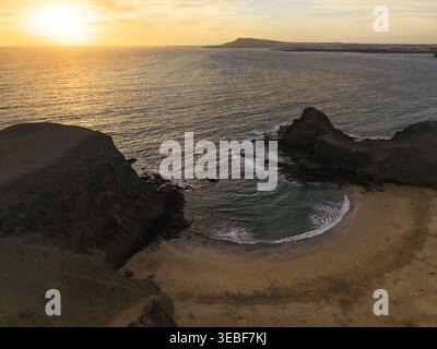 Vista aerea di una piccola insenatura di sabbia circondata da scogliere vulcaniche scure e calme acque oceaniche sotto un tramonto dorato Lanzarote, Canarias, Spagna. Foto Stock