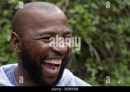 Uomo adulto afroamericano che ride con la bocca aperta e indossa una felpa con cappuccio grigio chiaro all'aperto con gli arbusti Foto Stock
