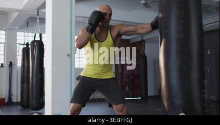 Pugni uomo muscoloso che colpisce la borsa pesante nella palestra di pugilato, indossa canotta gialla, pantaloncini grigi, guanti Foto Stock