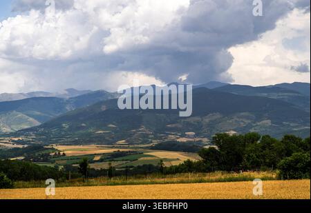 Ampio paesaggio rurale con campo dorato, colline verdi e lontane montagne blu sotto spettacolari nuvole di tempesta. Foto Stock