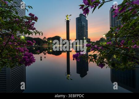 Didascalia: Vista all'alba del Monumento Selamat Datang a Bundaran HI, Giacarta, incorniciata da fiori di bouganville rosa e riflessa nell'acqua Foto Stock