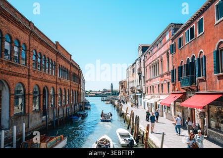 Venezia Italia - 25 aprile 2026: Canale di Venezia nell'isola di Murano con barche ed edifici storici, vivace strada sul lungomare e tradizionali archi italiani Foto Stock