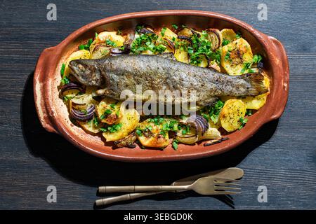 Trota intera al forno con patate e cipolla rossa in un piatto di ceramica. Trota arcobaleno tostata servita su un letto di patate al forno e cipolle con lei fresca Foto Stock