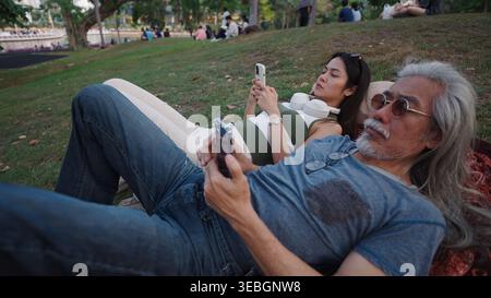 Un uomo anziano e una giovane donna sono sdraiati sull'erba in un parco, entrambi invischiati nei loro smartphone Foto Stock