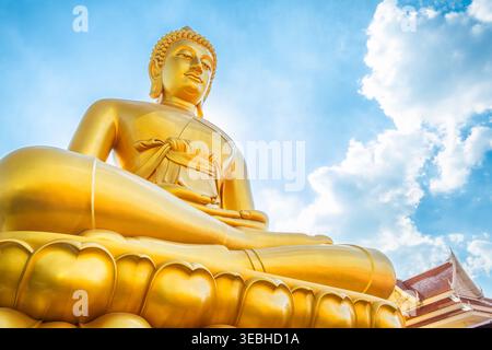 Gigante statua dorata del buddha, seduta su una base di loto contro il cielo blu con nuvole al Wat Paknam Phasi Charoen nella città di Bangkok, Thailandia. Vista ad angolo basso di Foto Stock