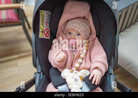 Simpatico baby sitter su un seggiolino per auto che indossa un vestito e un cappello rosa tenui, tenendo in mano un giocattolo. Concetto di infanzia, sicurezza e stile di vita familiare con calore e intimità Foto Stock