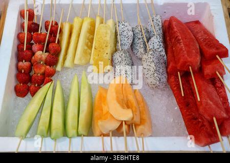 Spiedini di frutta tropicale assortiti su ghiaccio in un mercato tailandese di strada. Fragole fresche, ananas e bacchette di anguria in un espositore di schiuma. Foto Stock