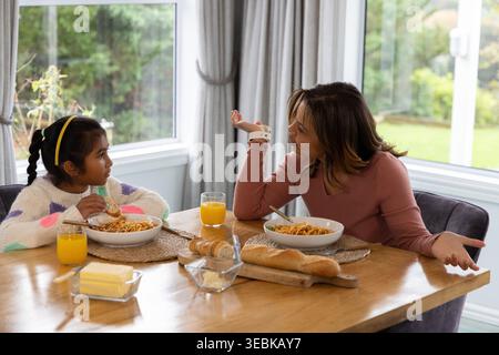 Donna di mezza età con top rosa e ragazza indiana con maglione seduto a casa a condividere la pasta Foto Stock