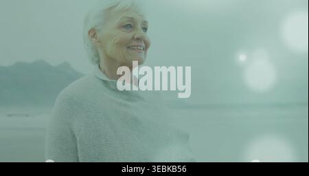 Donna anziana in piedi che indossa uno scialle in maglia grigia sul litorale, un bagliore calmo delle lenti marine, spazio per le copie Foto Stock