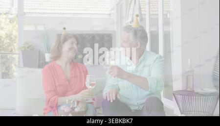 Brinda la coppia con il top rosso, camicia blu che solleva bicchieri da vino sul balcone con cupcake e cappelli da festa Foto Stock
