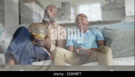 Padre e figlio indossano abiti casual che guardano la televisione nel soggiorno, tenendo in mano patatine e telecomando Foto Stock
