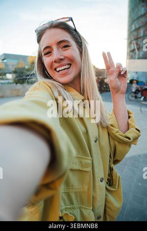 Una giovane donna allegra si fa felicemente un selfie e mostra giocosamente un segno di pace con le dita in piena luce del sole, irradiando gioia e positività con Foto Stock