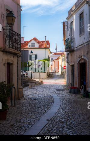 Lisbona, Portogallo - 7 maggio 2022: Vista di una tortuosa strada acciottolata soleggiata incorniciata da edifici storici con varie texture e roo con piastrelle rosse Foto Stock