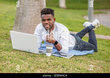 Vedi un afroamericano sdraiato sull'erba verde sorridere e pollice in su, coinvolto nel suo lavoro su un portatile. Foto Stock