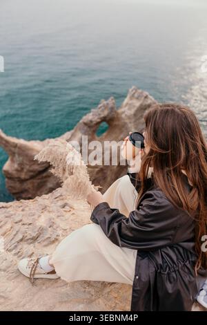 Donna che beve caffè nell'oceano seduto su una scogliera rocciosa bevendo da una tazza di carta e tenendo l'erba pampas durante una tranquilla mattinata Foto Stock