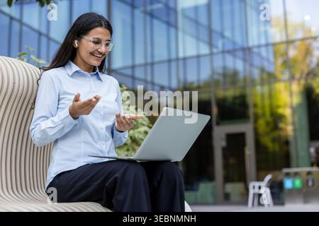 Giovane donna d'affari che indossa occhiali e auricolari wireless, comunica attivamente durante una videochiamata sul suo laptop mentre è seduta su una panchina moderna Foto Stock