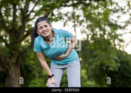 Donna in piedi all'aperto, che tiene lo stomaco e soffre di dolore, che raffigura intensi crampi addominali o mal di stomaco durante una sessione di attività fisica in un parco Foto Stock