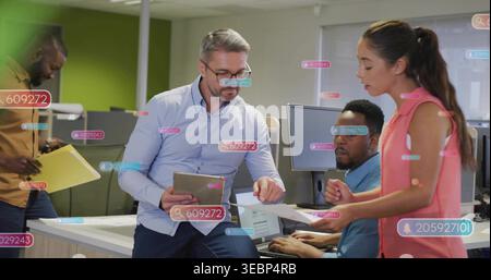 Punta l'uomo dai capelli grigi con una camicia blu in ufficio appoggiato alla scrivania con un tablet, mostrando carte e cartellini Foto Stock