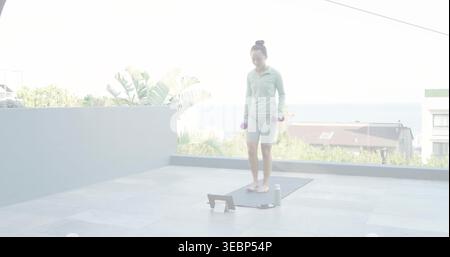 Donna che indossa un top verde e un paio di pantaloncini corti, tenendo i manubri viola sul tappetino del tetto con un tablet Foto Stock