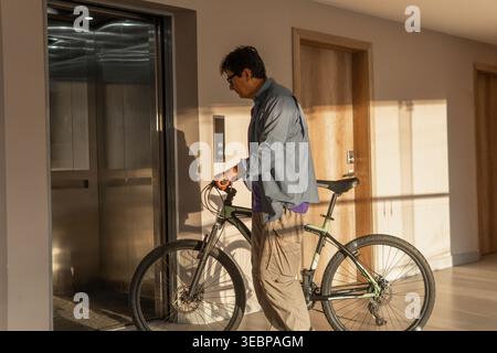 Uomo in possesso di una mountain bike, in attesa di un ascensore in un corridoio di appartamenti urbani Foto Stock