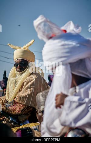 Zaria City, Nigeria - 22 marzo 2026: Vista di una figura regale con abiti tradizionali color crema calda e occhiali da sole scuri, un sorprendente contrasto con il bricco Foto Stock