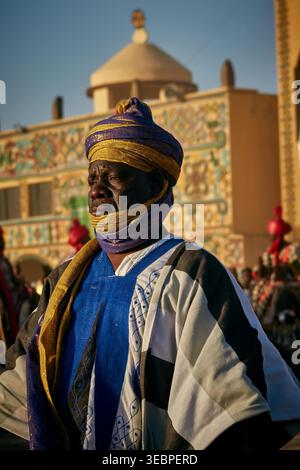 Zaria City, Nigeria - 22 marzo 2026: Vista di un uomo in un vivace abbigliamento tradizionale blu e giallo, con uno sguardo solenne, in piedi davanti a un intricato p Foto Stock
