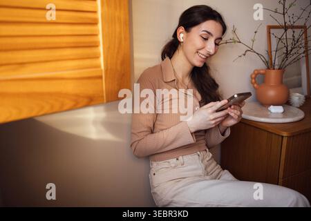Una giovane donna con lunghi capelli scuri ama usare il suo smartphone mentre è seduta contro una parete in un ambiente interno caldo e invitante con elementi in legno Foto Stock
