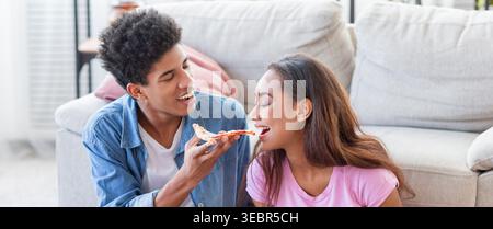 Giovani coppie che amano la pizza insieme sedendosi sul pavimento in un accogliente soggiorno Foto Stock