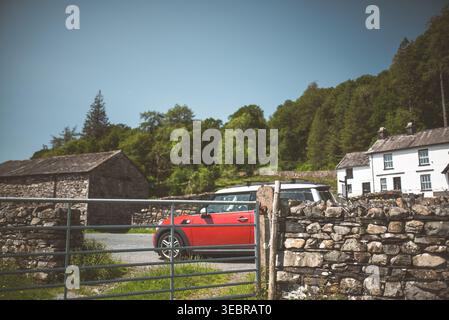 Una piccola auto rossa vibrante si trova parcheggiata vicino a un muro di pietra e ai tradizionali edifici inglesi in una giornata di sole Foto Stock