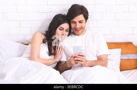 Coppia che si gode un momento di relax insieme mentre naviga su uno smartphone a letto Foto Stock