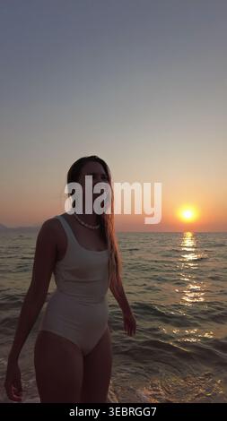 Donna in costume da bagno bianco che cammina sulla spiaggia fino al tramonto sul mare Foto Stock