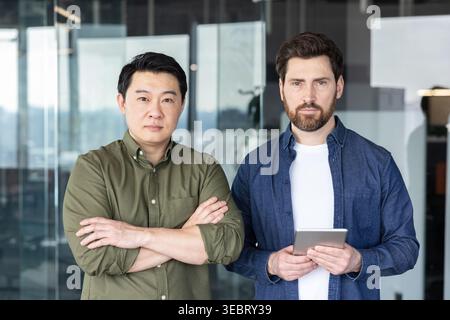 Due uomini d'affari fiduciosi e multirazziali si trovano in un moderno ufficio di startup, uno con le braccia incrociate, l'altro con un tablet che trasmette il lavoro di squadra, leader Foto Stock