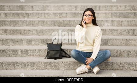 Donna seduta sui gradini impegnata in una conversazione telefonica mentre indossa un abbigliamento casual Foto Stock