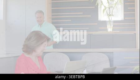 Lavoratrice anziana che scrive sul computer portatile al tavolo dell'angolo cottura, indossa camicetta di corallo, uomo che guarda Foto Stock