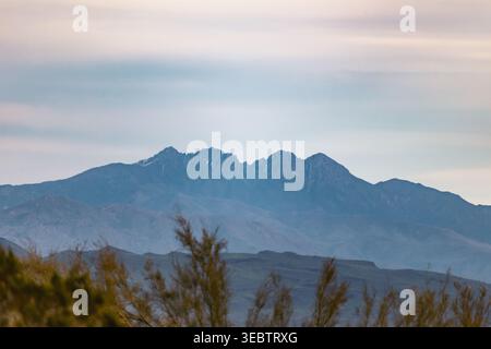 Catena montuosa lontana sotto un cielo morbido e nebuloso. I picchi sagomati si innalzano dietro le boccole in primo piano. La vegetazione sparsa incornicia il paesaggio aspro e nebbioso. MU Foto Stock