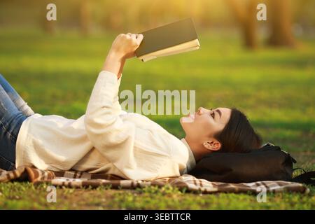 Leggere un libro mentre si è sdraiati sull'erba in un parco durante una giornata di sole Foto Stock