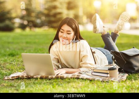 Giovane donna che si diverte a lavorare sul laptop nel parco durante il pomeriggio soleggiato mentre si rilassa sulla coperta Foto Stock