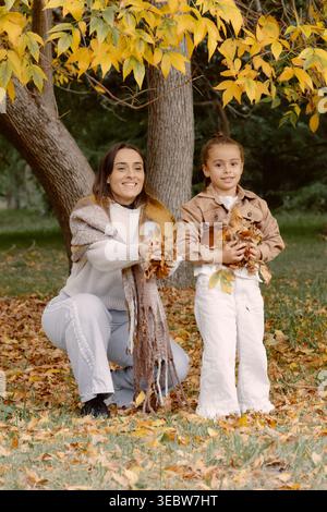 Mamma e figlia si godono l'autunno insieme, raccogliendo le foglie cadute sotto gli alberi dorati, condividendo un divertente momento all'aperto pieno di caldo clima stagionale Foto Stock