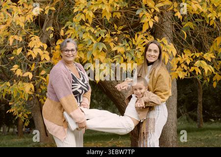 Nonna, madre e figlia giocano insieme in un parco autunnale, sollevando il bambino orizzontalmente sotto alberi dorati, catturando una gioia e un'energia Foto Stock