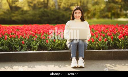 Giovane donna che lavora su un laptop nel parco circondata dai vibranti tulipani rossi sotto il cielo soleggiato Foto Stock