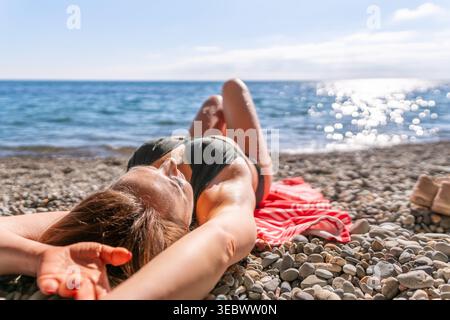 Donna che prende il sole sulla spiaggia sdraiata sulla spiaggia di ciottoli vicino all'oceano blu durante le vacanze estive Foto Stock
