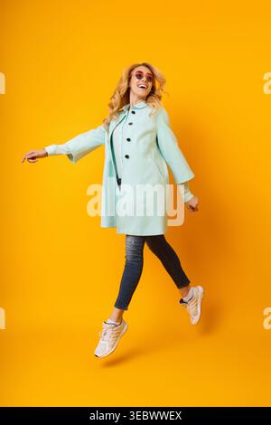 Una giovane donna salta con gioia nell'elegante cappotto Mint su un vivace sfondo giallo Foto Stock